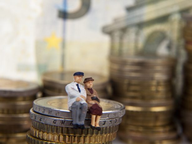 Kaksi miniatyyrihahmoa, siististi pukeutuneet vanhempi nainen ja mies, istuvat kolikkopinon päällä.